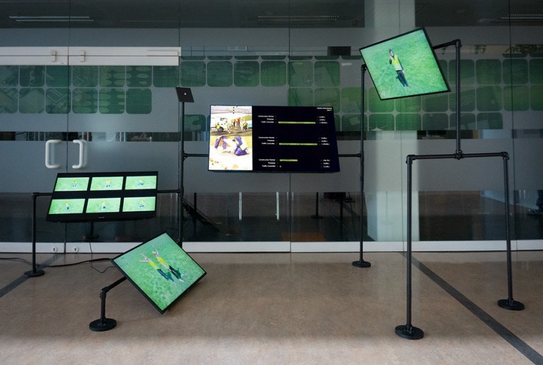 Vision Processor: EN471 (2019), video installation, Vision Processor chip, machine learning, video dataset, found footage, Rotterdam, The Netherlands Installation in an exhibition space. Made from black scaffolding tubes at different heights with a screen on each support. The three smaller outside screens show video’s of archetypal physical behavior for protestors, construction workers and traffic controllers wearing yellow vests. They are surrounded by grass. While the center screen shows existing video footage of protestors and the classification percentages from the machine learning algorithm.